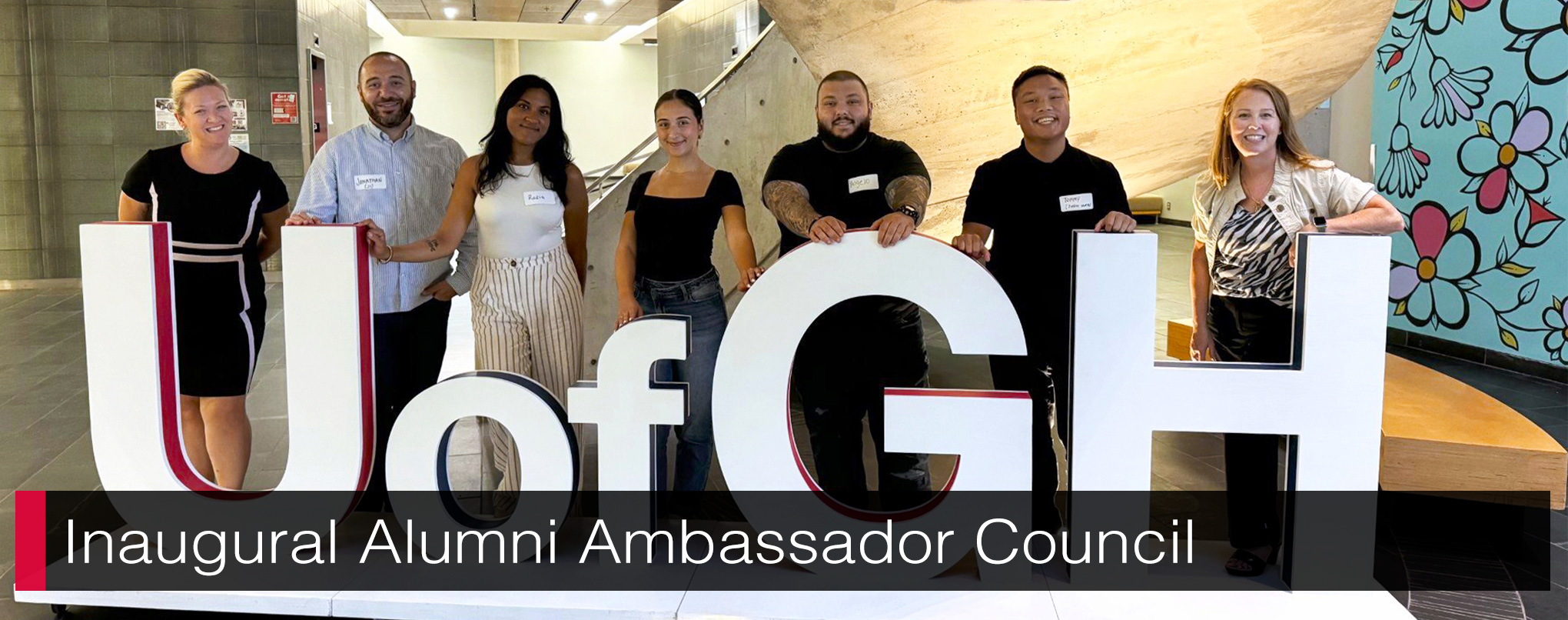 Seven individuals standing behind large, white UofGH letter with text, 