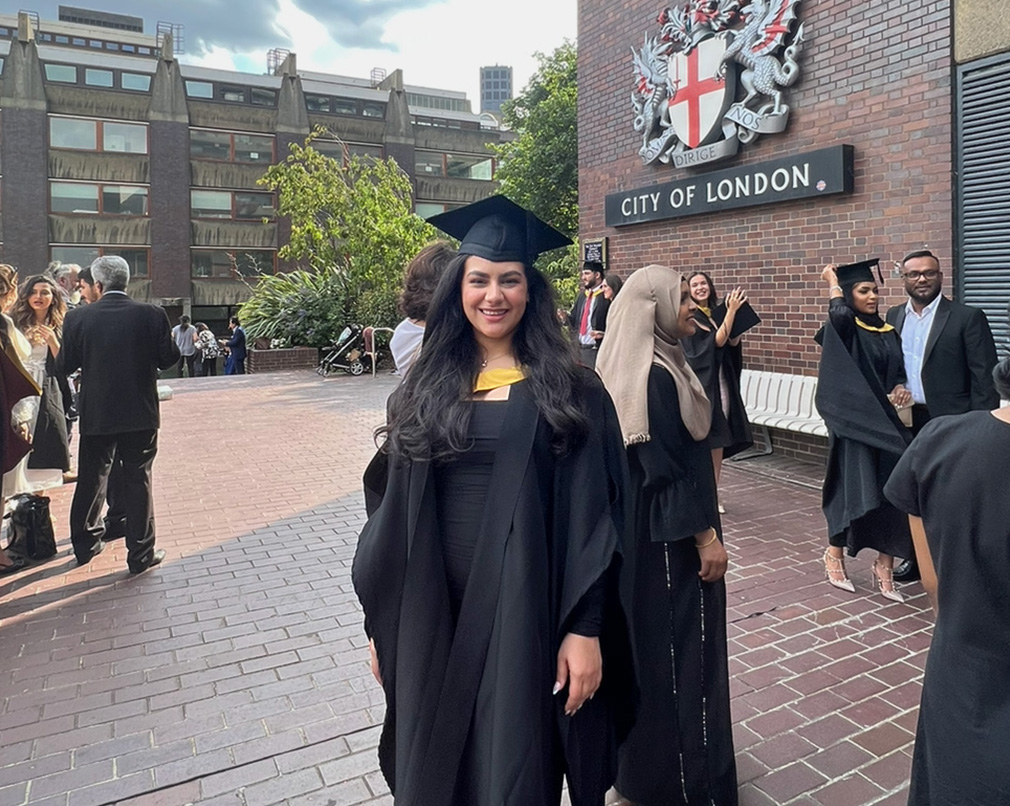 Someone in graduation regalia standing in front of City of London crest in the background