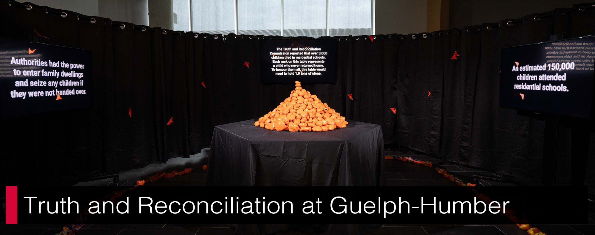 A neatly pile of orange-painted rocks sit on a table with three TV screens with the surrounding.