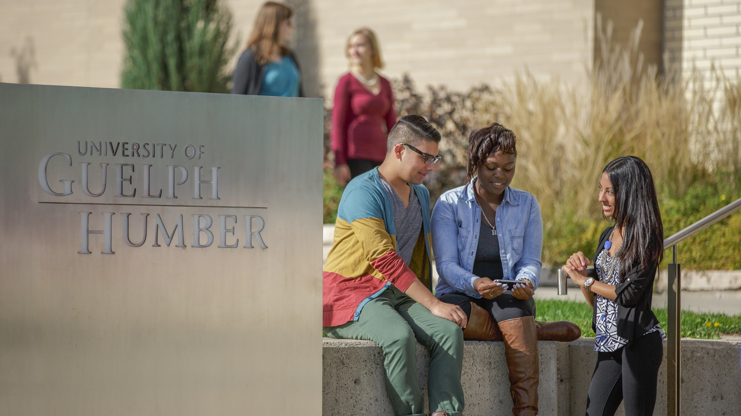 Three students sitting on ledge outside of the GH building next to the sign that says University of Guelph-Humber